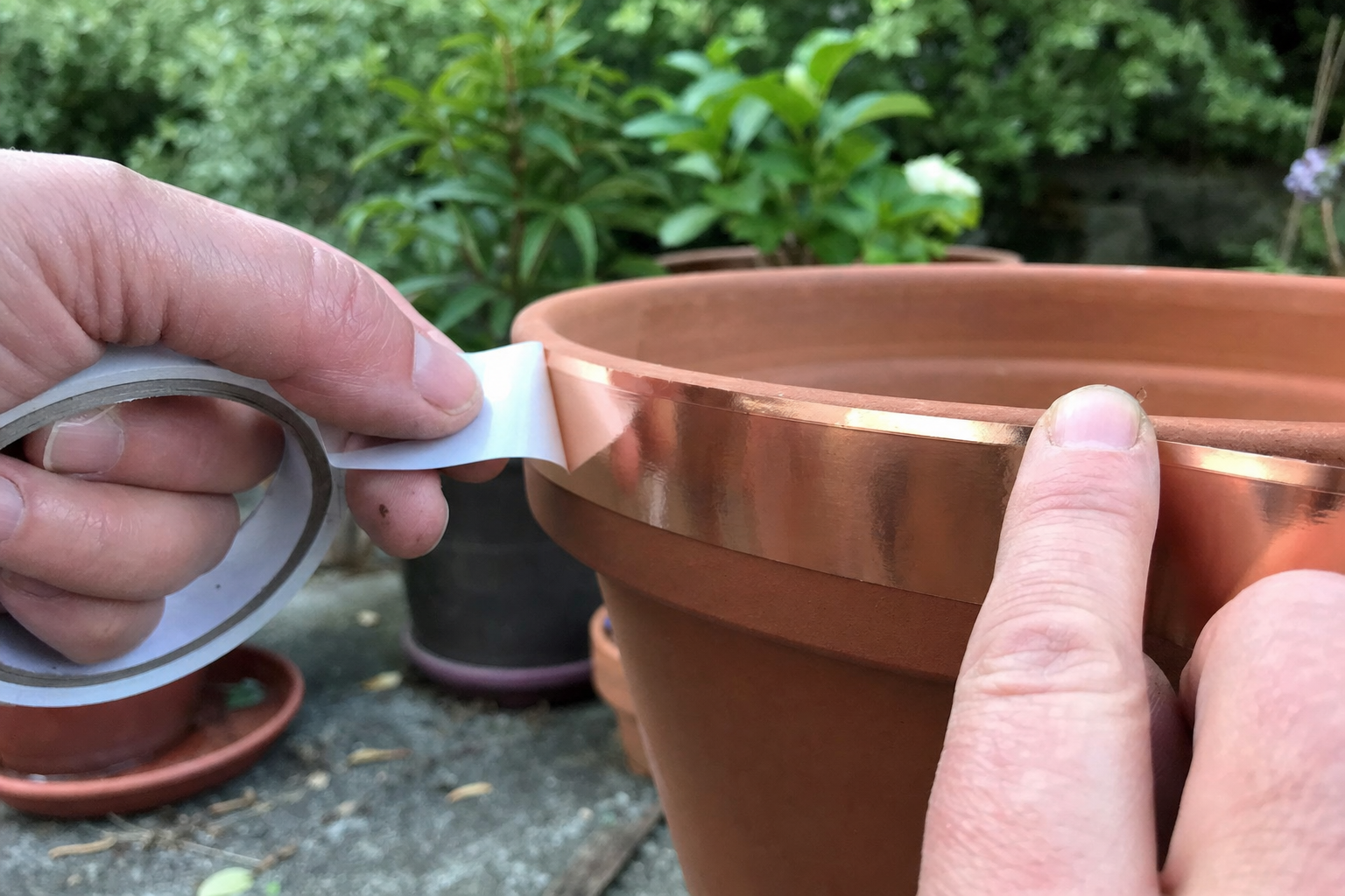 Hands peeling the backing and pressing bright copper tape firmly onto a clean terracotta pot rim in a garden